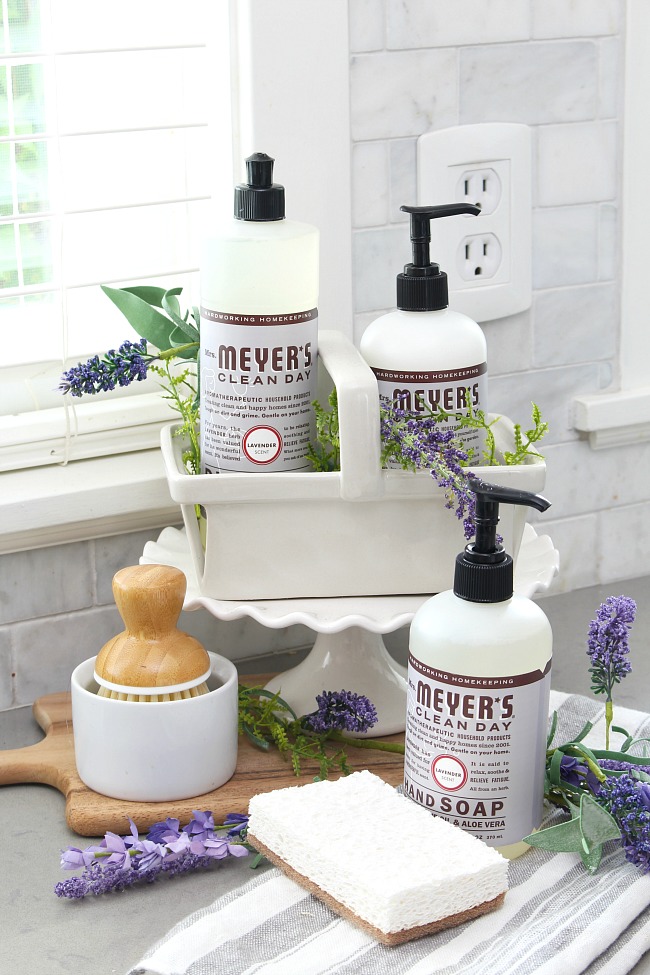 Mrs. Meyers lavender dish soap, hand soap, and hand lotion displayed on a cake stand by the kitchen sink.
