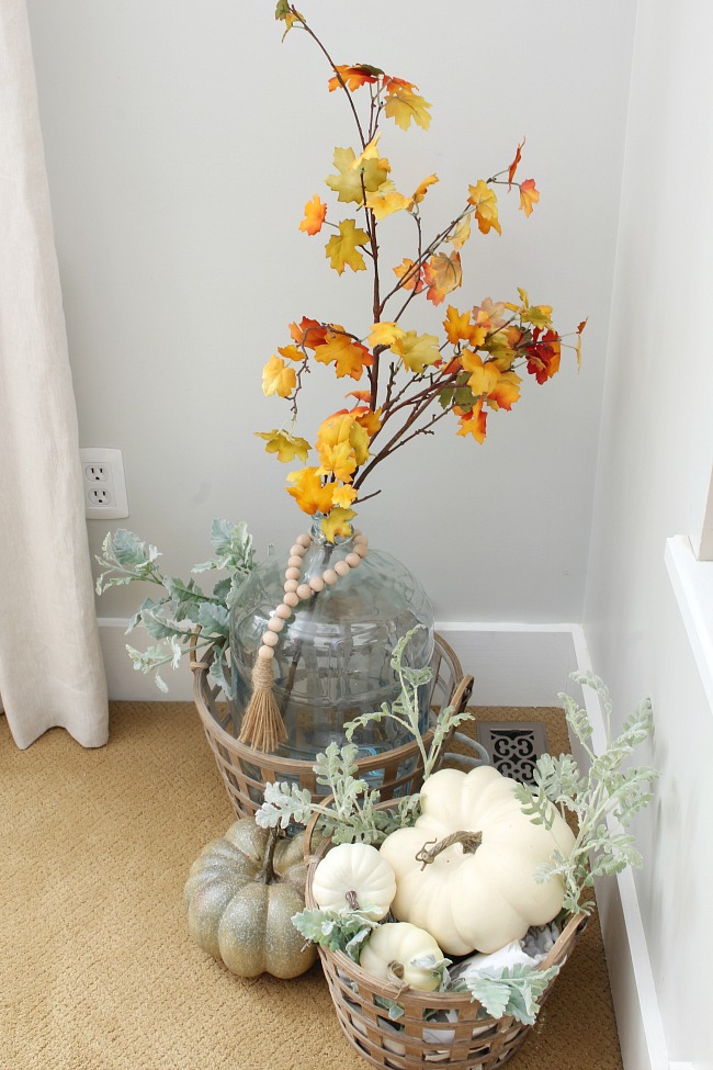 Demijohn with fall colored leaves and neutral pumpkins in a fall basket.