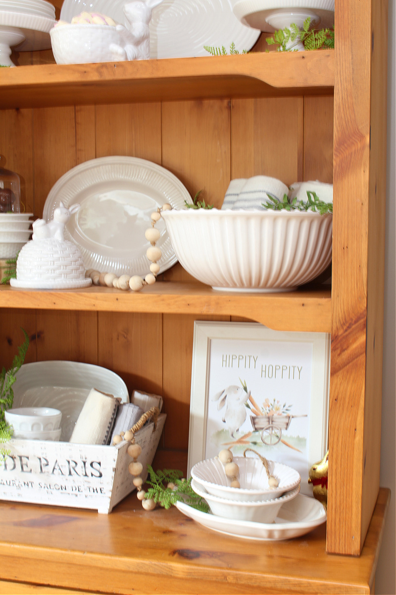 Wood hutch decorated for Easter with white dishware, greenery, and a few Easter touches.