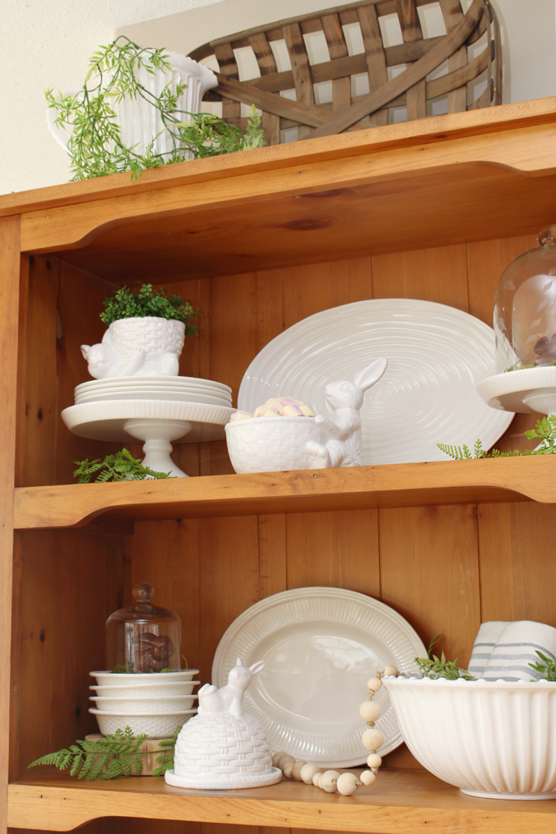 Wood hutch decorated for Easter with white dishware, greenery, and Easter bunnies.