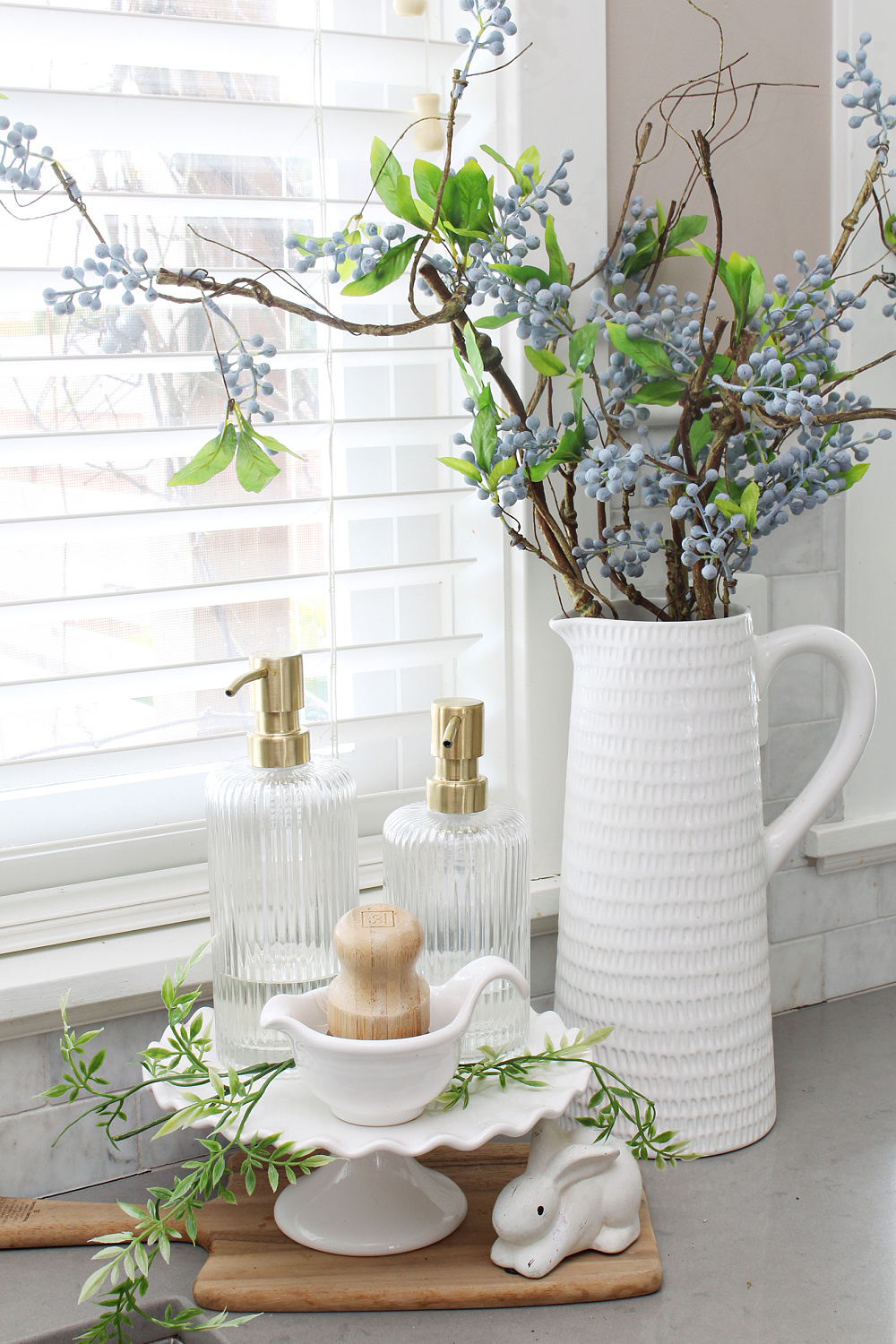 Sink cleaning supplies on a white cake stand with white vase filled with faux branches. 